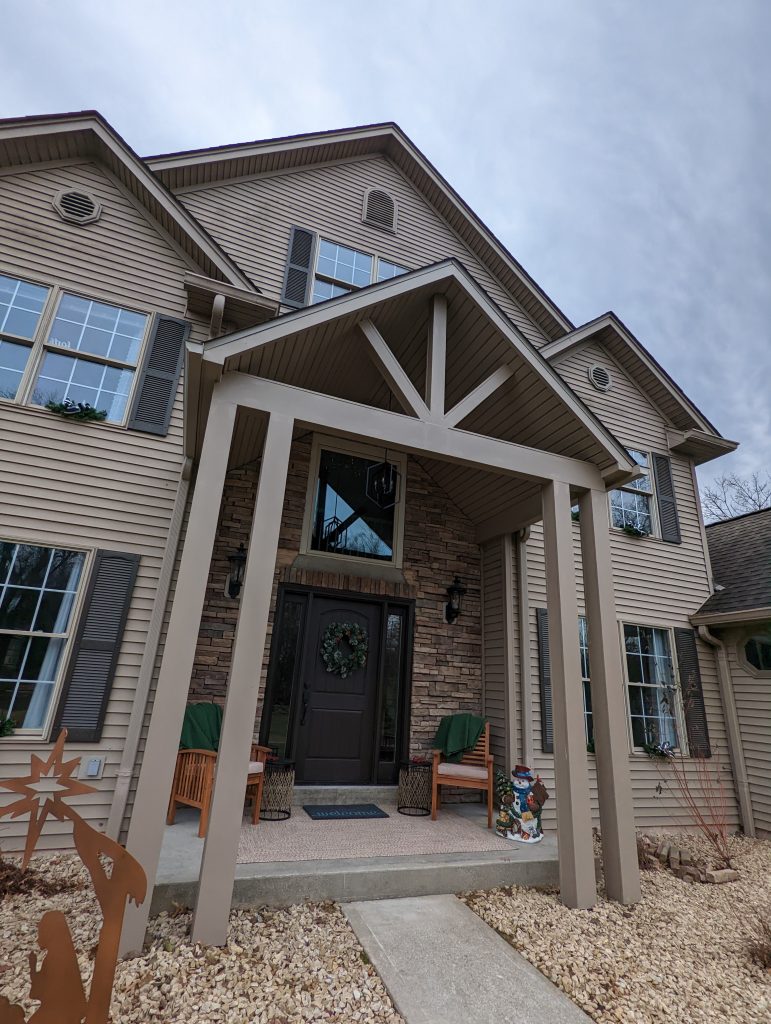 Exterior view of a two-story home featuring beige siding, a stacked stone entryway, and a custom covered front porch by Mueller Remodeling.
